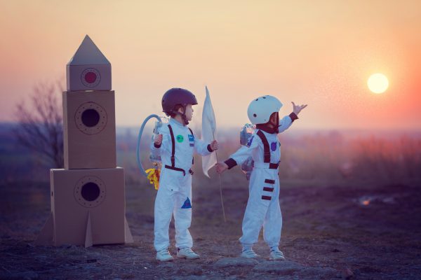 Two kids in Astronaut costumes standing next to cardboard rocket ship holding flag and pointing toward the setting sun